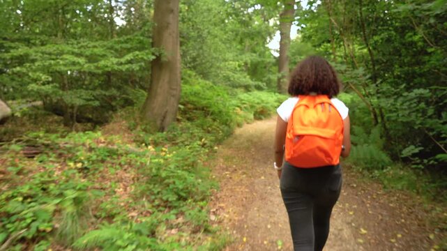 Teenage mixed race African American girl young woman hiking with orange backpack, smart watch and take out coffee cup in forest woodland 