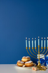 A vertical festive Hanukkah scene featuring a gold menorah with lit candles, a plate of powdered...