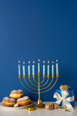 Naklejka na ściany i meble A vertical festive Hanukkah scene with a lit menorah, soft candle glow, donuts and coins, and wrapped gifts on a blue backdrop. Warm, celebratory mood suitable for holiday greetings