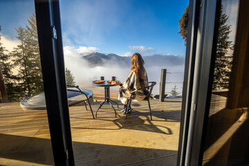 Woman eating breakfast on mountain cabin terrace. Woman wrapped in a blanket enjoys breakfast on a wooden terrace, gazing at misty mountain peaks under a clear blue sky.