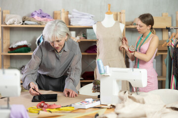 Mature dressmaker draws a design on an electronic tablet against the background of an assistant in a sewing workshop.