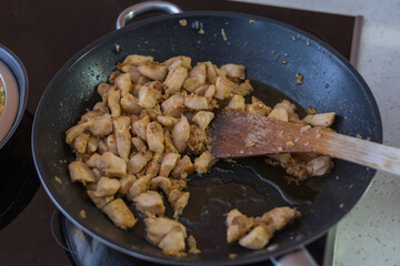A top-down view of a black non-stick frying pan on a stovetop, containing diced chicken pieces being fried.