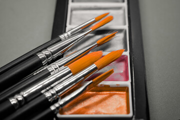 Close-up of artist paintbrushes resting on a watercolor palette showing bright orange bristles and metallic ferrules in a creative studio atmosphere with soft light and detailed texture