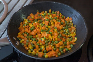 A top-down view of a black non-stick frying pan on a stovetop, containing a brightly colored vegetable mix being fried