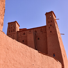 Red clay walls and towers rise high into a clear sky in Ait ben haddou, Morocco. Ancient earthen construction and architecture are seen on a sunny day
