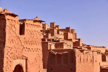 This is a view of the ancient Ksar of Ait ben Haddou. The traditional mud brick buildings are stacked on a hillside with a clear blue sky above, showing the texture of the architecture