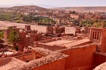 This shows an aerial view of the historic Ksar of Ait Benhaddou in Morocco. A village sits beyond it. It is during the day, and the landscape is bathed in a warm, desert light.