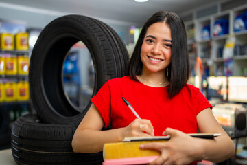 Smiling woman writing on clipboard, holding air filter and tire, in auto service shop