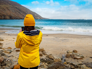 Girl in yellow jacket and hat is looking at sandy beach, ocean, cliffs and beautiful blue cloudy sky. Travel and tourism. Amazing Keem beach and bay in county Mayo, Ireland. Sightseeing nature.
