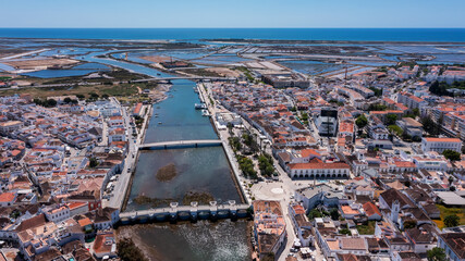 Aerial view of Tavira with Roman bridge over Gilao River traditional whitewashed buildings salt pans and Atlantic Ocean in the distance under clear sky in Algarve. Concept of historical coastal charm