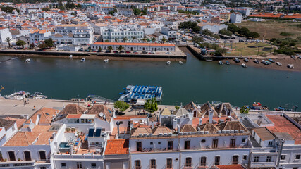 Aerial view of Tavira town with traditional tiled rooftops and boats along the Gilao River under bright sunlight in Algarve, Portugal. Concept of historic riverside tourism