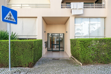 The entrance of a contemporary apartment building is framed by well-maintained hedges and a pedestrian crossing sign indicating the safe walking area