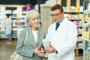 In pharmacy, man apothecary holds package with therapeutic cream for sensitive skin in hands and talks with senior woman client about nuances of care.