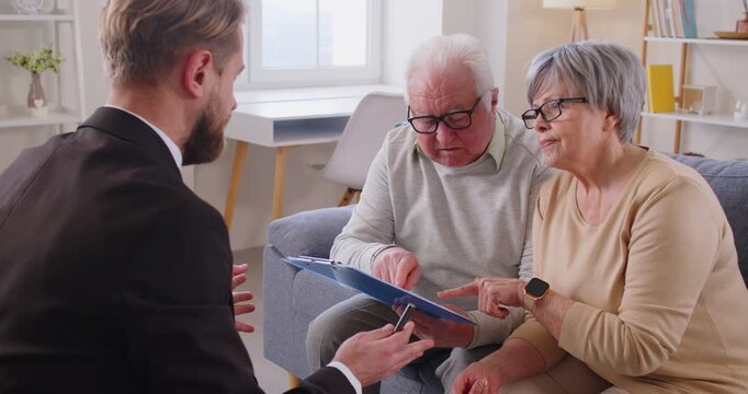 Elderly spouses sitting on couch attentively review agreement documents, listening to explanations of young advisor, agent, lawyer or insurer. Retirement planning, investment advice, finances, will