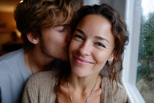 This delightful image captures a couple sharing tender moments while embracing the view from a rain-streaked window, radiating warmth and happiness during cozy times together.