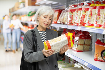 Mature female in shop carefully examines information about product on label packaging. Elderly woman customer chooses and buys pack of Chinese noodles at hypermarket