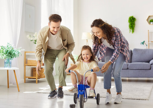 Happy family parents and daughter play at home riding bicycle. In livingroom, child girl rides tricycle in helmet as dad guides and mom cheers. Fun, safety, supportive parenting and leisure concept.