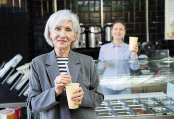 Smiling senior lady standing beside bubble tea display, savoring taste of popular Taiwanese refreshing beverage with chewy tapioca boba in disposable cup