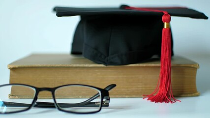 Close up of a graduation cap atop a tan book with a red tassel and black framed glasses set against a stark white background, a classic still life academic study.