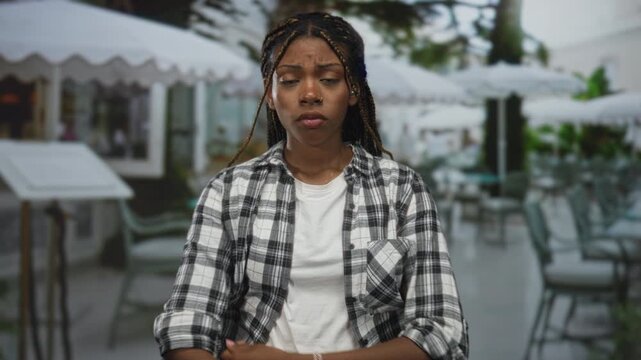 Woman with arms crossed on outdoor restaurant terrace wearing plaid shirt and white tee, braided hair and stern expression; frustration refusal.