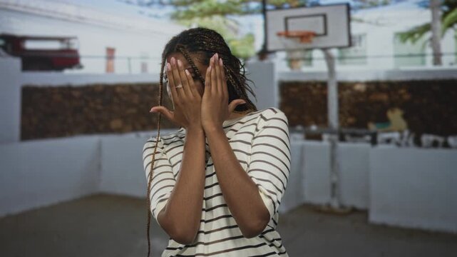 Woman covering face with hands, braided hair and visible ring on finger, striped shirt on street basketball court; shyness privacy.