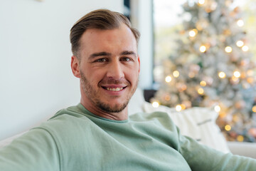 Mid-adult man relaxing on light sofa at home, gazing toward window showing decorated Christmas tree
