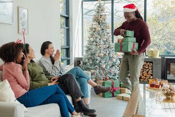 Diverse friends clapping on sofa at home receiving gifts from man in Santa hat, copy space