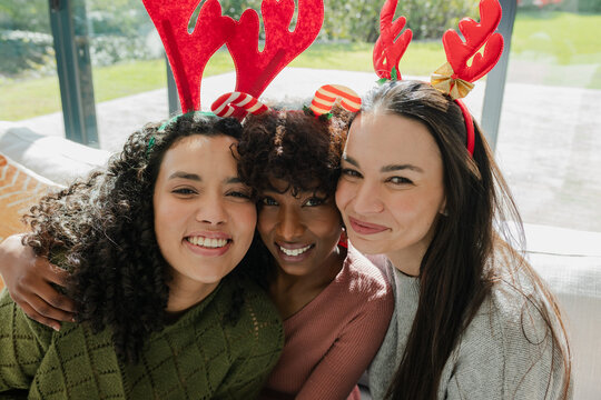 Diverse female friends laughing and leaning on sofa in living room wearing red antler headbands
