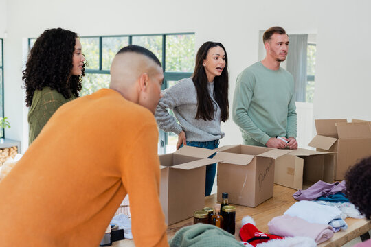 Diverse volunteers sorting and labeling boxes with clothing and jars on wooden table in bright room