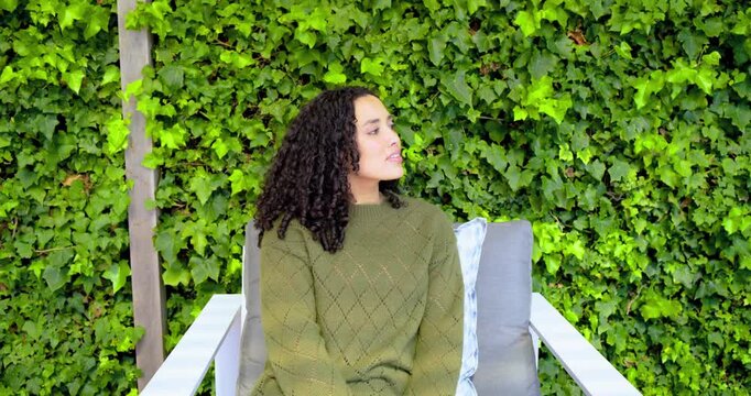 Hearing rustle, curly-haired woman sitting on patio chair shifting gaze across ivy wall and smiling