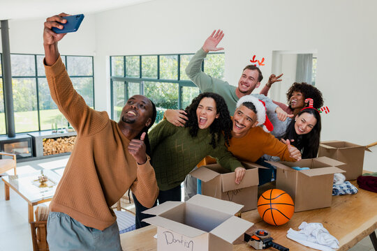 Diverse friends wearing festive hats unpacking boxes holding smartphone in living room cheering
