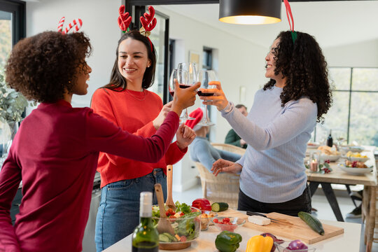 Diverse female friends raising wine glasses at kitchen island wearing antler headbands, wine bottle - Powered by Adobe