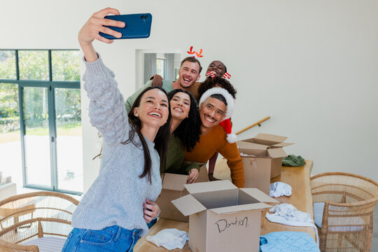 Diverse volunteers taking selfie while sorting donation boxes and clothing at wooden dining table
