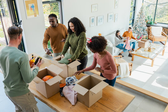 Diverse friends packing sweaters and bottles into boxes around table at home by Christmas tree
