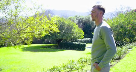 Man entering sunlit garden, crossing arms, turning toward camera, greeting with warm smile