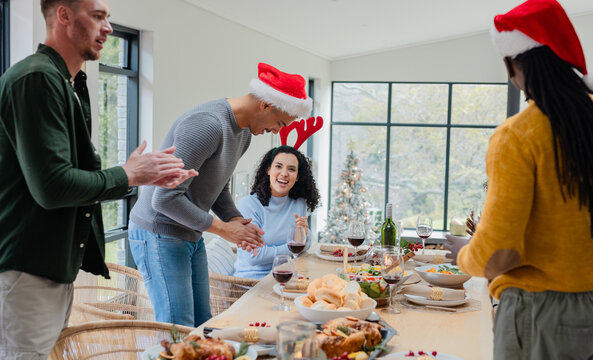 Diverse friends wearing Santa hats gathering around wooden table in dining room with Christmas tree