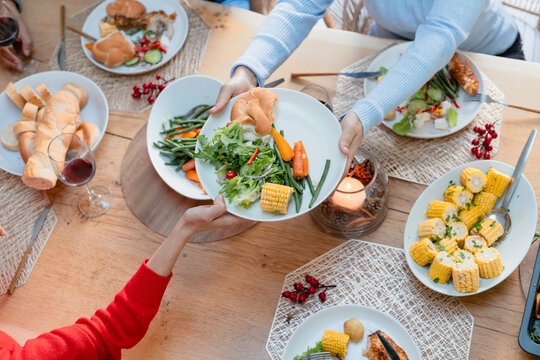 Diverse mother and daughter passing plates around dining table at home with salad plates, candle