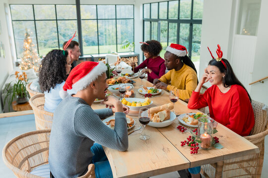 Diverse friends wearing festive hats gathering at home by table drinking wine and eating turkey