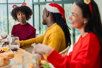Diverse friends wearing festive headwear sharing meal at wooden table at home with wine glasses
