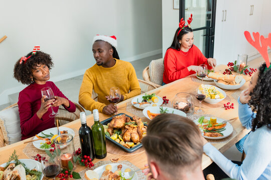 Diverse friends wearing sweaters holding wine glasses sharing roasted chickens around dining table
