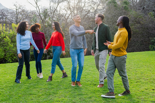 Diverse friends walking and chatting across grassy lawn in park with leafless trees and shrubs