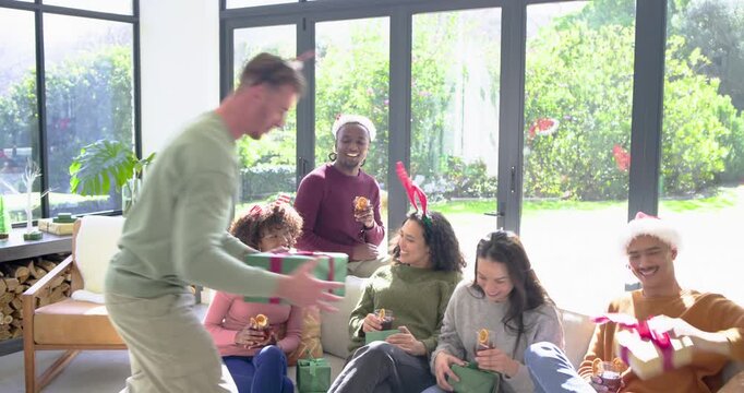 Amid eager diverse friends, mid adult white man entering home carrying wrapped gifts, handing them