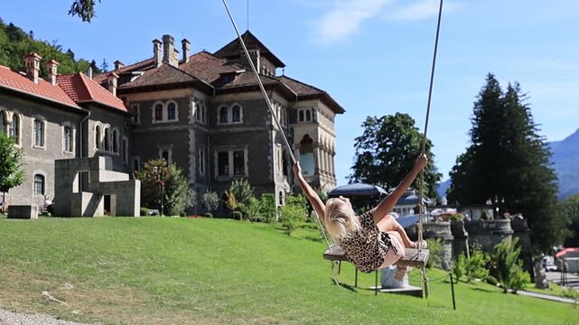 Woman playing on a swing, enjoying the summer day with a historic castle and green mountains of Transylvania, showing leisure, happiness, and relaxed travel in Romania, Cantacuzino Castle of Busteni