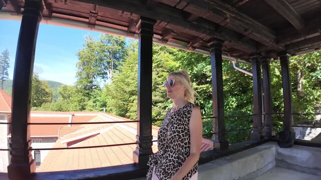 Woman smiling, looking away from a Cantacuzino castle balcony, enjoying the beautiful mountain landscape and clear blue sky while on vacation in Transylvania, Romania
