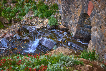 Mountain Waterfall Flowing Over Rocks With Stone Bridge And Wildflowers