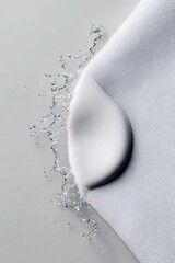A smooth white cream swatch is captured in motion with a dynamic splash of water and tiny golden beads against a textured white fabric.