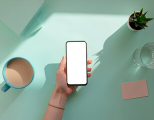 Overhead view of a hand holding a blank white screen smartphone with a cup of coffee and a plant on a light blue background