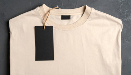 Plain beige t-shirt with a blank black tag, displayed flat on a dark grey textured surface
