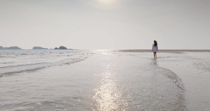 Woman walking along ocean shore on wide, expansive sand beach at sunset, soft waves lapping the waterline and distant islands on horizon, Ko Lanta, Thailand. People enjoy freedom, travel and relax