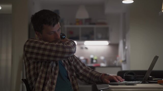 Cervical neck ache, nerve arthritis. Man studying late through night at laptop computer with cervical pain. Exhausted guy relaxing his neck using gun massager while overworking at notebook computer. 